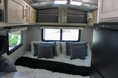 Cozy RV rental bedroom featuring a plush bed with white and gray pillows, overhead storage, and windows with outdoor views.