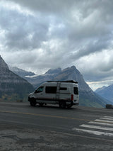 White RV rental van on a mountain road amidst majestic, cloud-covered, snow-capped peaks.
