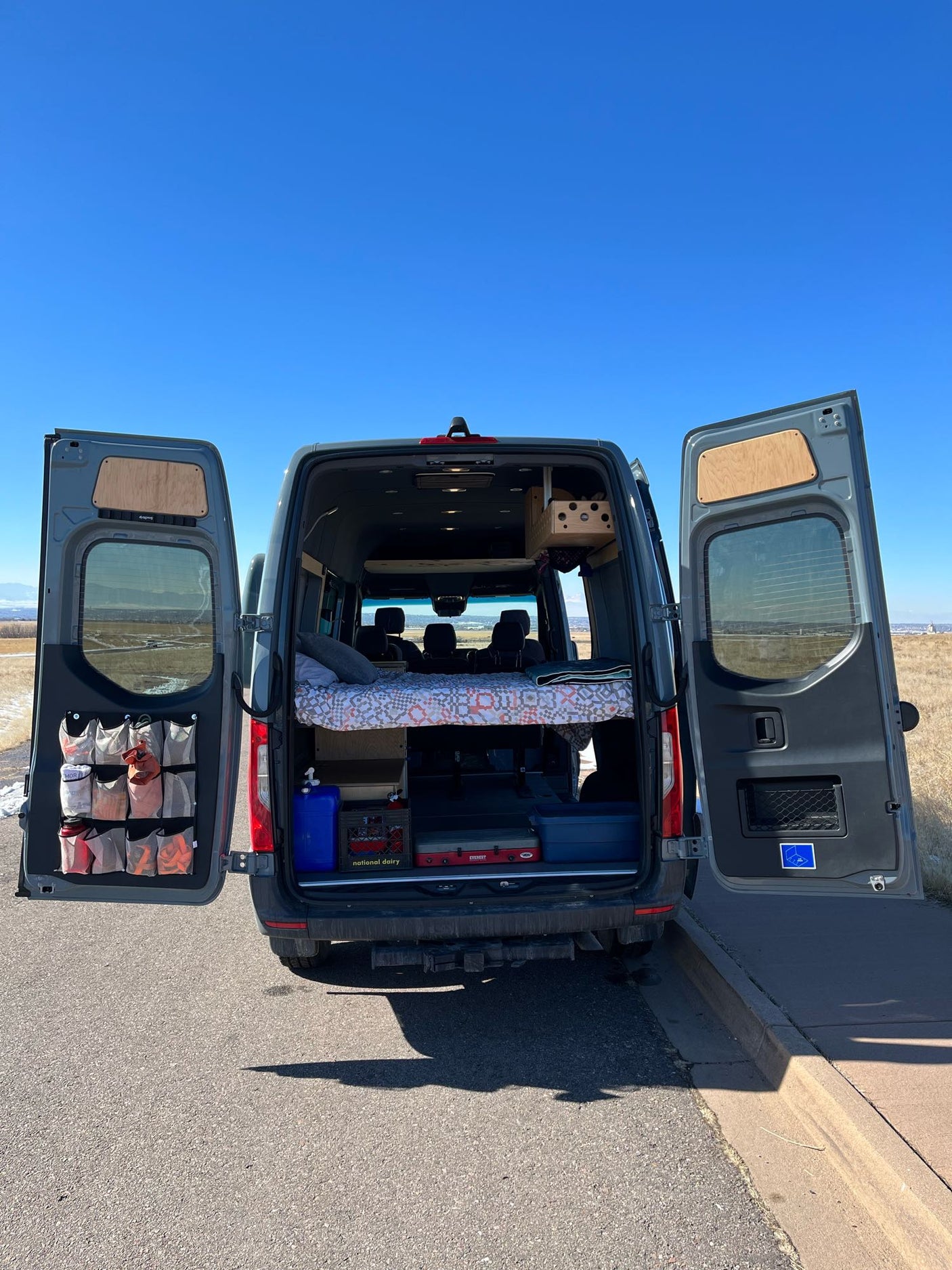 Interior view of an RV rental camper van with open back doors, showing a bed, kitchen gear & storage in a vast landscape.