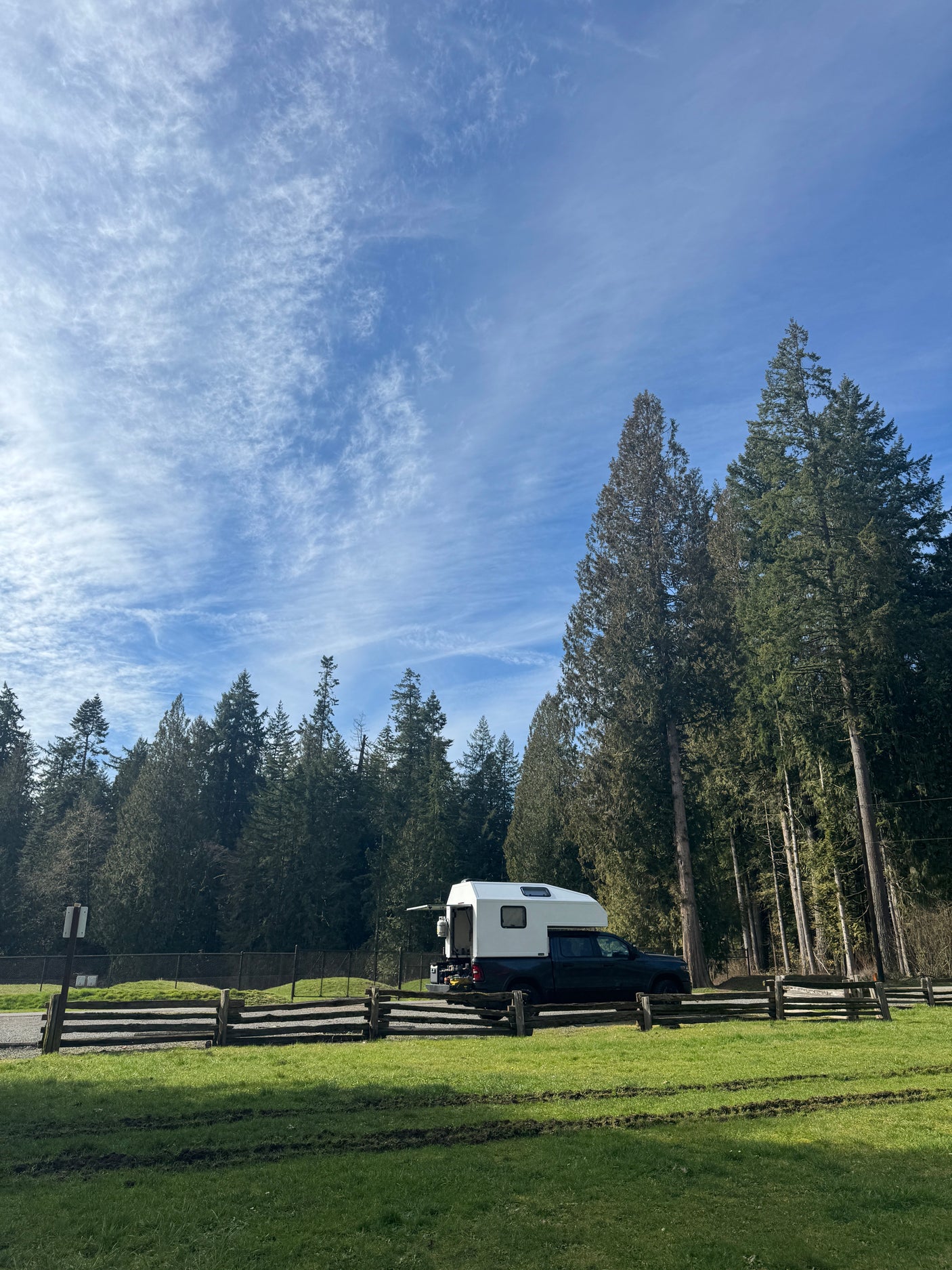 RV rental truck camper in a grassy spot by a wooden fence, backed by tall evergreen trees under a blue, cloudy sky.