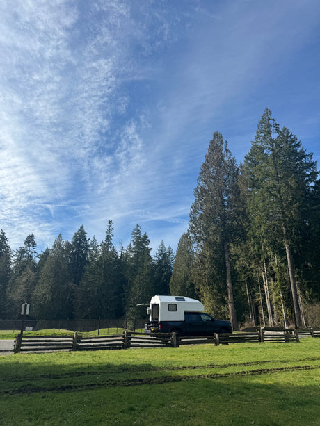 RV rental truck camper in a grassy spot by a wooden fence, backed by tall evergreen trees under a blue, cloudy sky.
