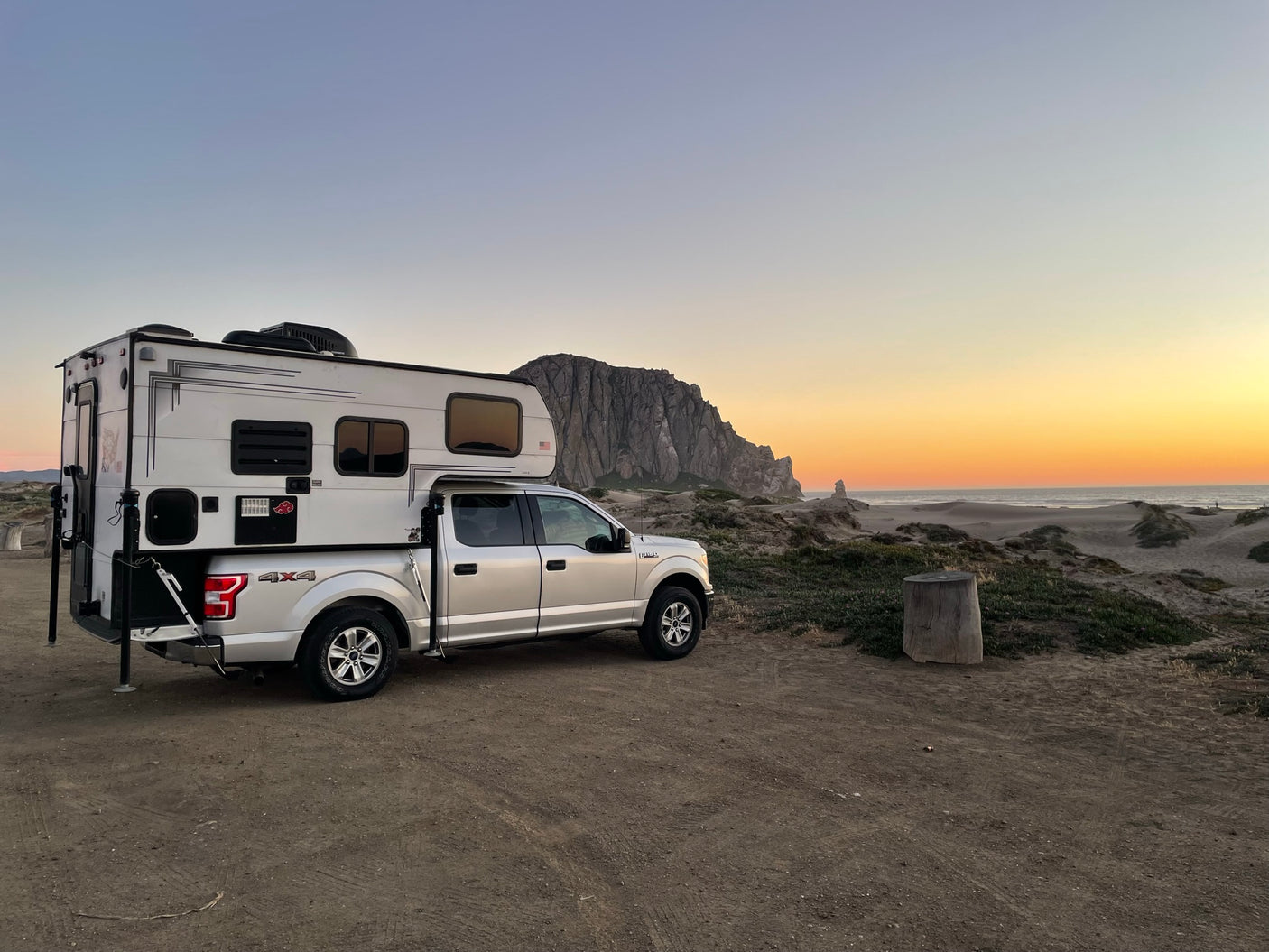 A white RV rental truck camper is parked on dirt near sandy dunes & Morro Rock at sunset by the ocean, ready for coastal adventures.