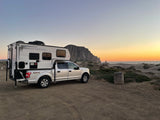 A white RV rental truck camper is parked on dirt near sandy dunes & Morro Rock at sunset by the ocean, ready for coastal adventures.