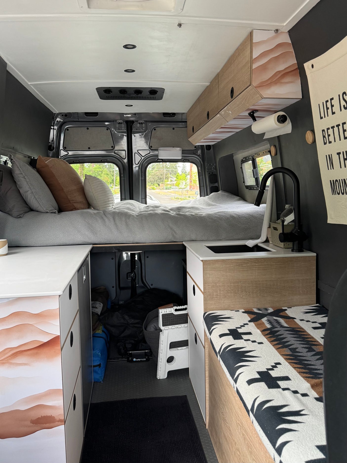 Inside a modern rv rental camper van, featuring a high bed, compact kitchen with sink, overhead cabinets, and under-bed storage.