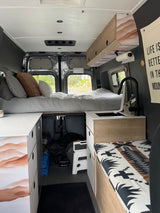 Inside a modern rv rental camper van, featuring a high bed, compact kitchen with sink, overhead cabinets, and under-bed storage.