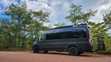 Dark grey RV rental with bikes on back, parked on dirt road in green forest. Perfect for nature adventures.