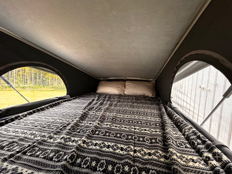 Experience comfort in this RV rental! A pop-top camper bed with a patterned blanket, pillows, and views of nature.