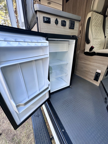 RV rental interior: open mini-fridge/freezer, power panel with outlets, and passenger seat. Compact and functional space.