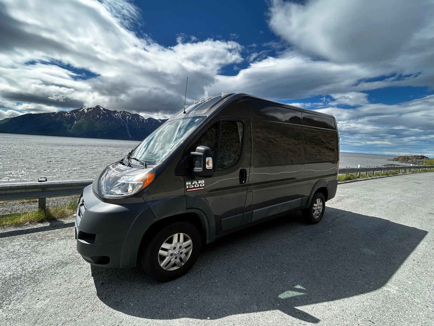 Dark grey Ram ProMaster RV rental parked on a scenic road by mountains and water under a cloudy sky, ideal for adventure.