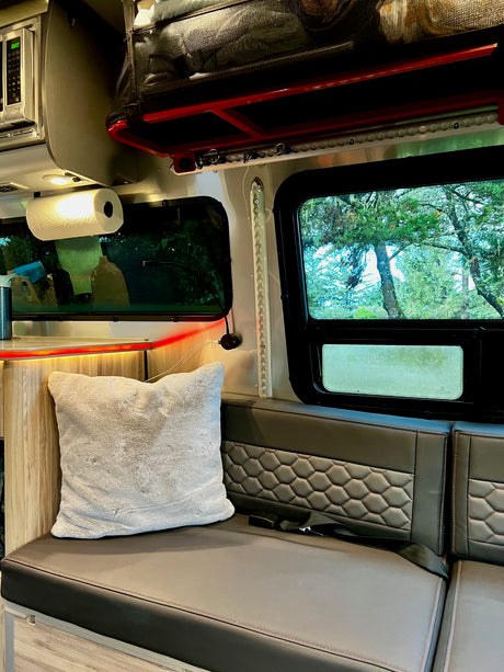 This cozy RV rental interior features a comfy bench with a plush pillow, microwave, and scenic window view of trees.