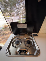 Inside an **rv rental**, a two-burner gas stove (Suburban brand) with lighting instructions and a screened window view of trees.