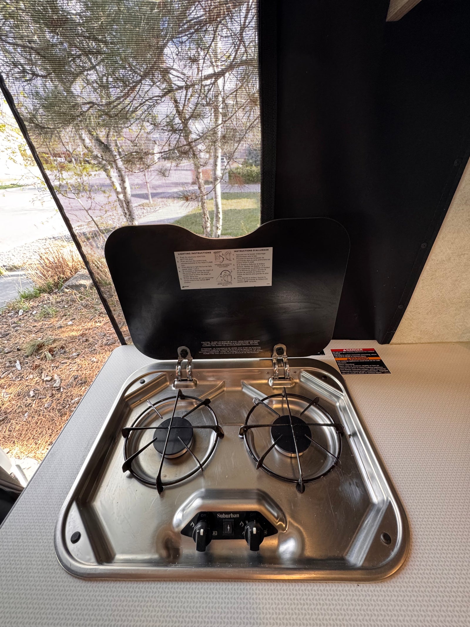 Inside an **rv rental**, a two-burner gas stove (Suburban brand) with lighting instructions and a screened window view of trees.