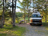 RV rental parked at a serene, wooded campsite by a lake, featuring a purple picnic table and leveling blocks under the van.