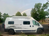 A double rainbow arches over a white rv rental camper van parked on a residential street with trees.