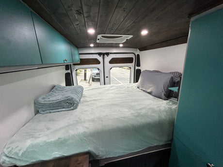 Inside a cozy rv rental camper van: large bed with teal bedding, overhead cabinets, wood plank ceiling, and open rear doors.