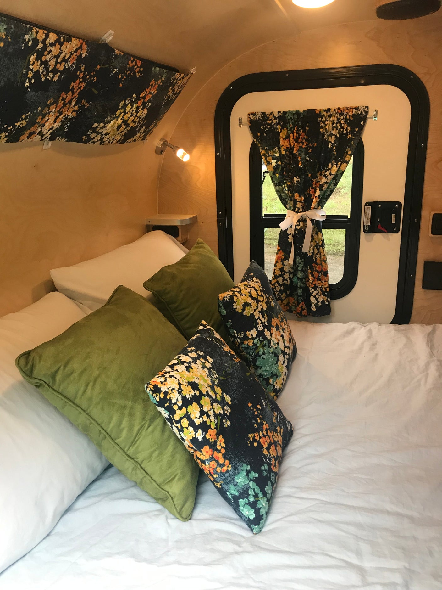 Cozy interior of an RV rental: a bed with white sheets, green pillows, and floral accent pillows, next to a window with a matching floral curtain.