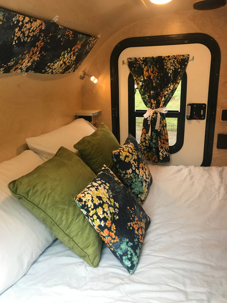 Cozy interior of an RV rental: a bed with white sheets, green pillows, and floral accent pillows, next to a window with a matching floral curtain.