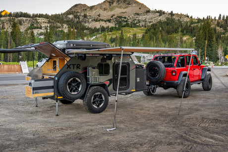 This rv rental setup features a red Jeep and a grey overlanding trailer with an extended awning and kitchen, ready for mountain camping.