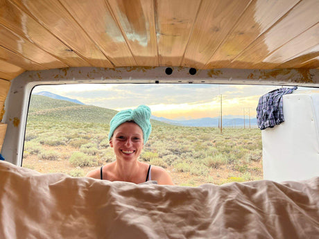 RV rental adventure! A smiling woman with a towel-wrapped head looks out from her van at a serene desert sunset.