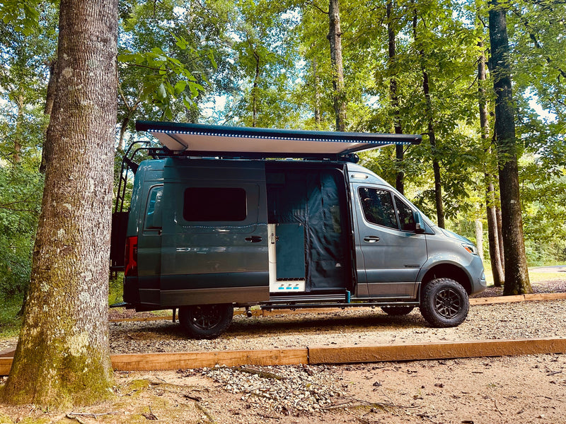 Your next outdoor adventure starts with this Winnebago RV rental. A grey camper van with an illuminated awning, parked in a wooded campsite. — photo 2