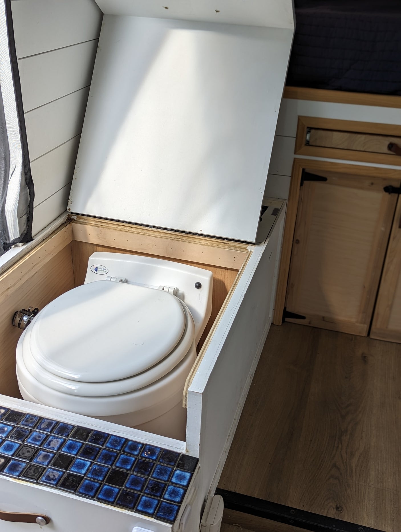 Compact RV rental toilet setup: a white portable toilet in a wooden enclosure with a lift-top lid and blue accent tiles.