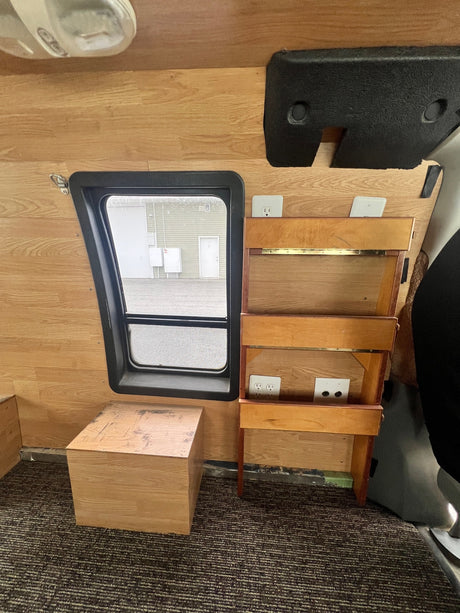 RV rental interior with wood paneling, carpeted floor, window, and custom shelf unit featuring power outlets.