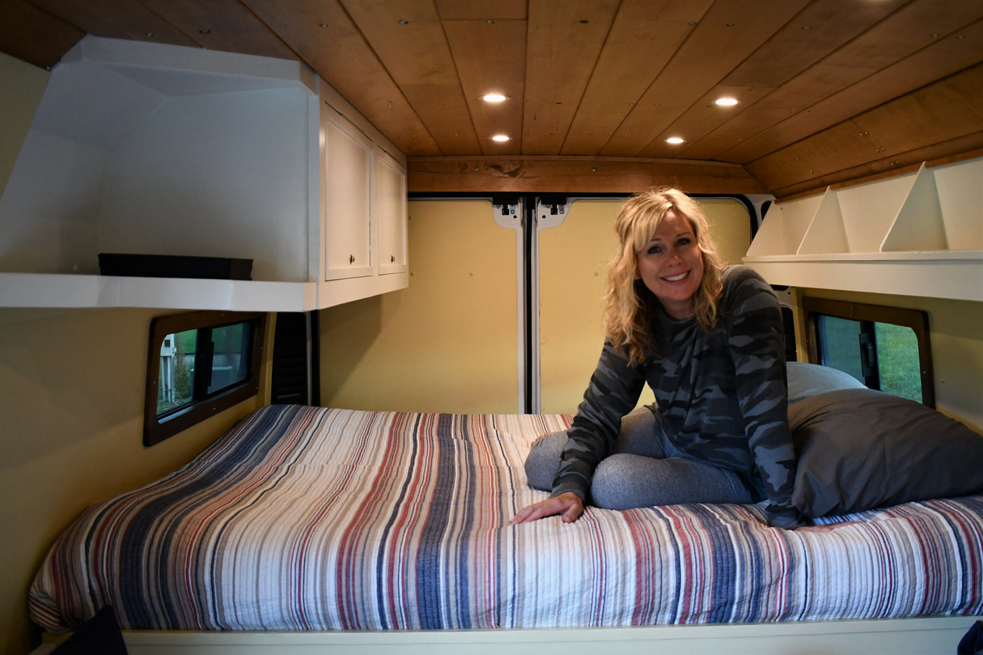 Smiling woman on striped bed in RV rental. Cozy van interior with wood ceiling, built-in cabinets, and rear van doors.