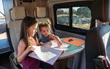 Two girls learn in an RV rental. They are seated at a table inside a camper van, sharing a large activity book.