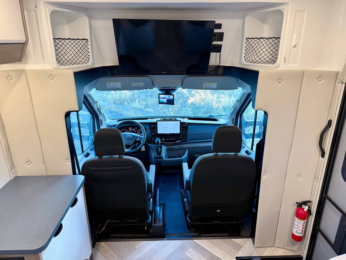 Interior view of an RV rental looking towards the cockpit with the driver's area, dashboard, and an overhead TV. A fire extinguisher is visible.