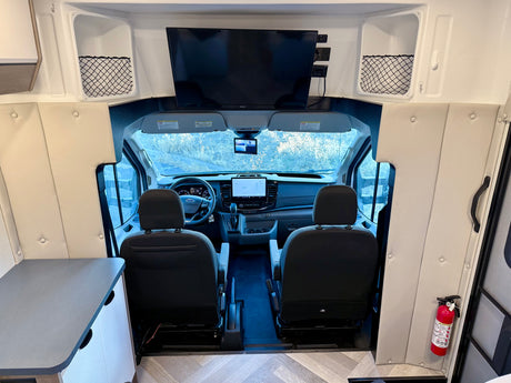 Interior view of an RV rental looking towards the cockpit with the driver's area, dashboard, and an overhead TV. A fire extinguisher is visible.