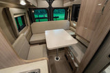 U-shaped dinette inside a modern RV rental: light cushions, table, and large windows revealing a vibrant green outdoor view.