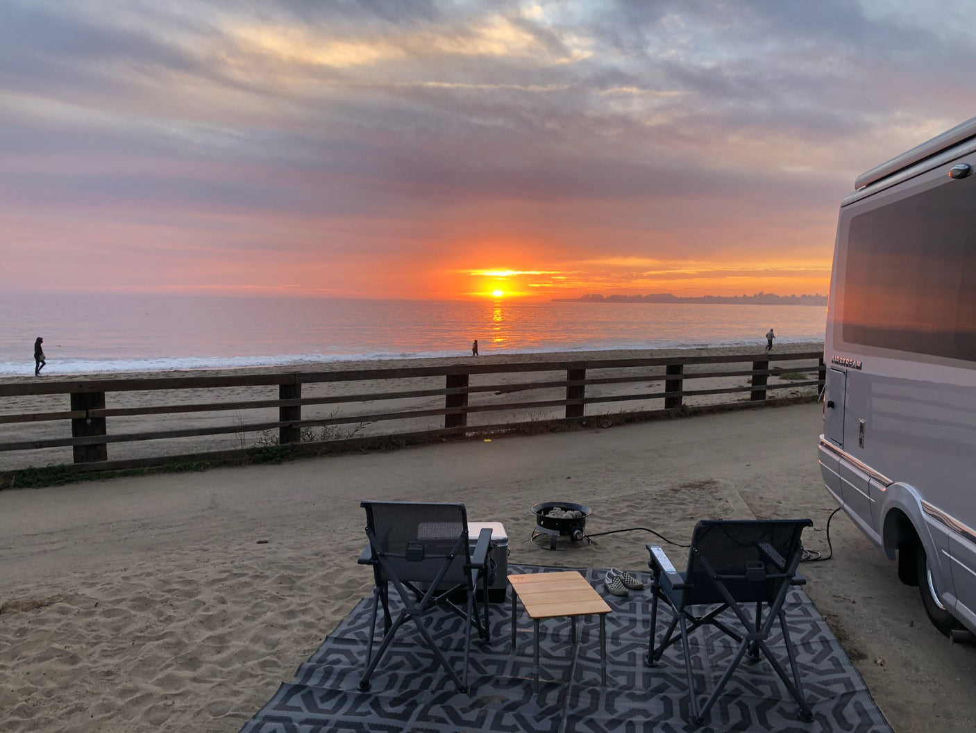 Enjoying a beautiful beach sunset from an RV rental campsite. Two chairs, a fire pit, and people walking by the ocean. Ideal for travel.