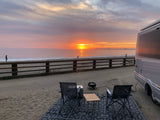 Enjoying a beautiful beach sunset from an RV rental campsite. Two chairs, a fire pit, and people walking by the ocean. Ideal for travel.