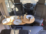 Interior of an RV rental, featuring swiveling front seats, dashboard, and a wooden dining table set with plates and cups. Diamond plate floor.