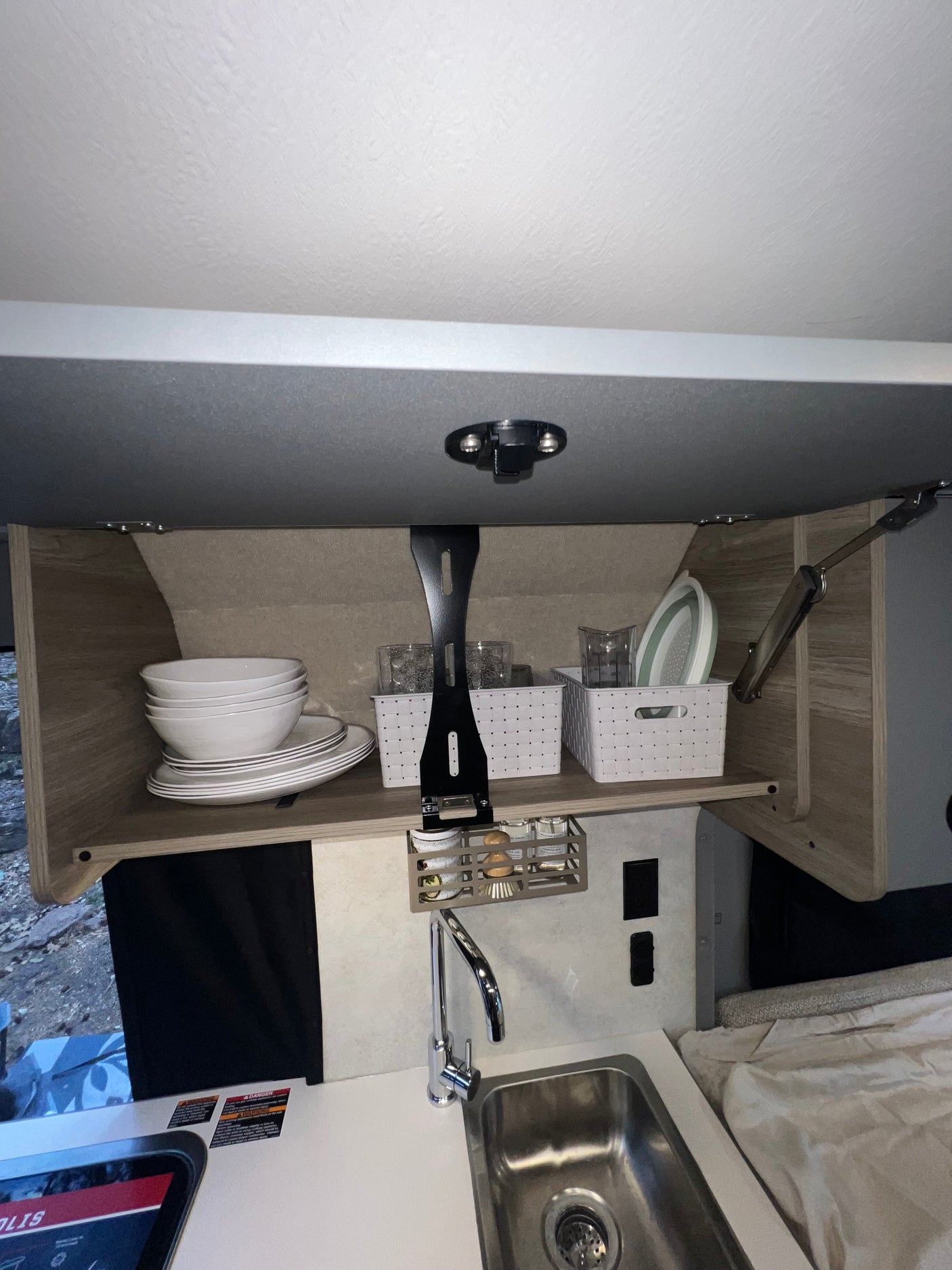 An RV rental's kitchen galley: open overhead cabinet filled with dishes, glasses, and baskets, above a sink with a faucet.