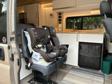 Inside an RV rental: A car seat is secured on a bench, facing a kitchenette with a sink and refrigerator, ready for family travel.