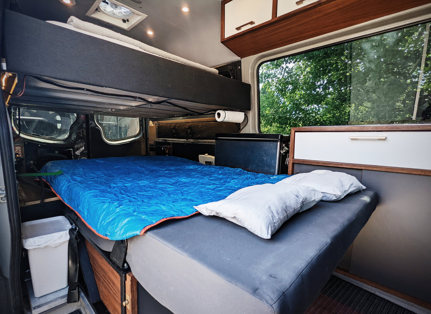 This RV rental offers a spacious interior: main bed with blue sleeping bag, an upper bunk, and ample storage.