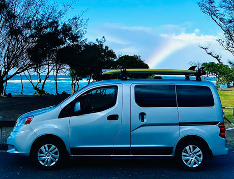 Scenic rv rental parked by the ocean with a surfboard on top & rainbow. Perfect for coastal adventures.