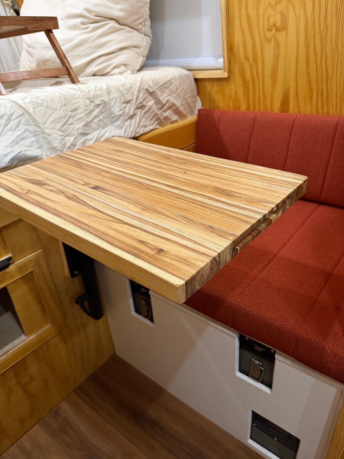 Cozy RV rental interior featuring a light wood table with a unique grain, a red upholstered bench, and a comfortable bed. — photo 13