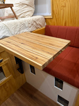 Cozy RV rental interior featuring a light wood table with a unique grain, a red upholstered bench, and a comfortable bed.