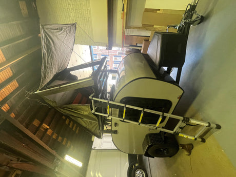 This RV rental features a compact travel trailer with a deployed rooftop tent, parked inside a garage.