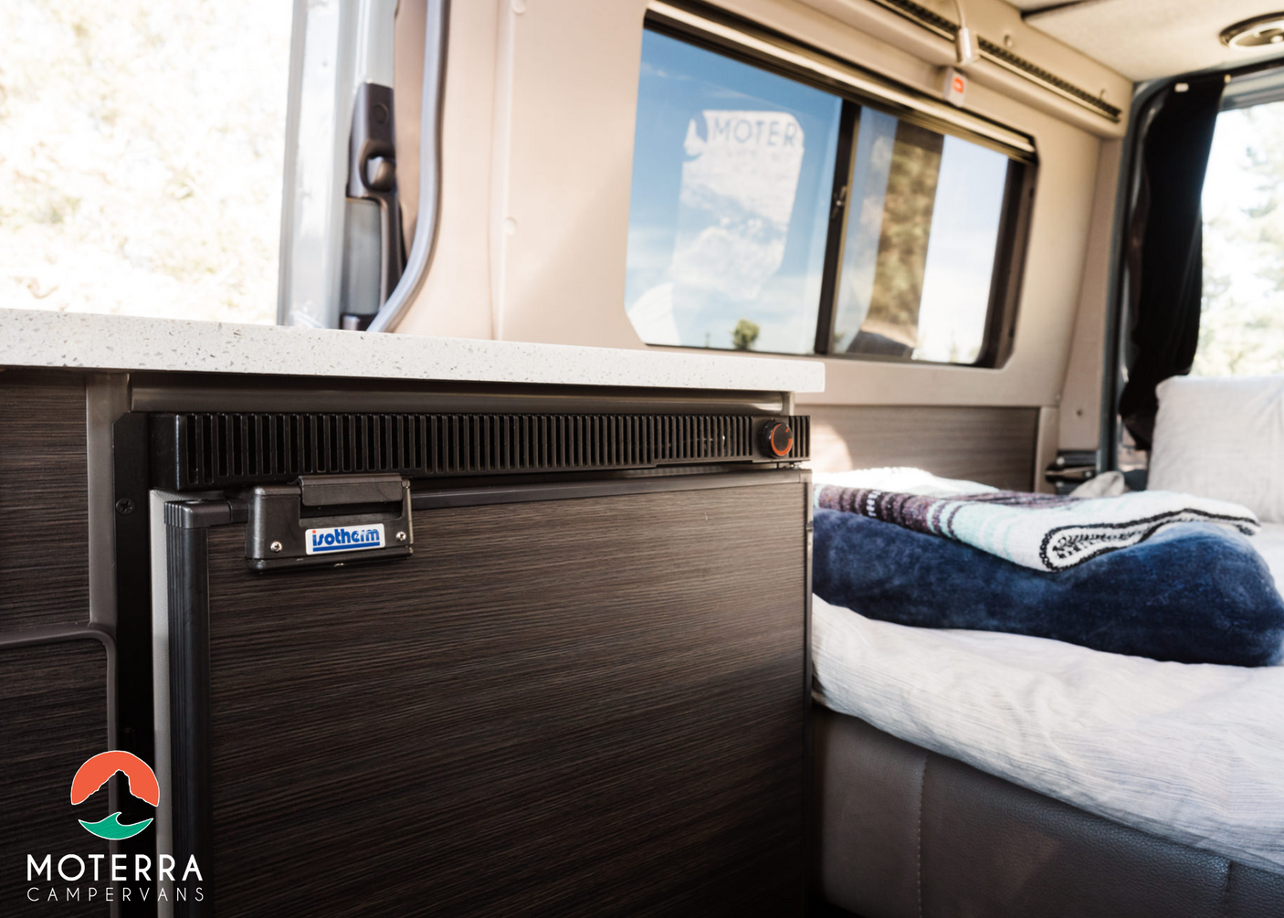Experience comfort in this Moterra rv rental: a built-in fridge, cozy bed, and countertop in a modern camper van interior.