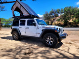 A white Jeep Wrangler Rubicon with an open Roofnest tent, parked on sandy ground. Ideal for your adventurous rv rental.