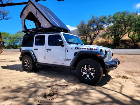 A white Jeep Wrangler Rubicon with an open Roofnest tent, parked on sandy ground. Ideal for your adventurous rv rental.