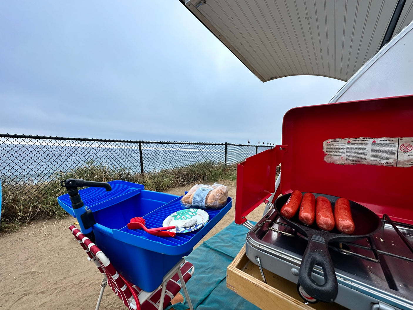 Outdoor cooking by the ocean: Hot dogs sizzle on a camp stove, set up outside an RV rental, offering scenic views.
