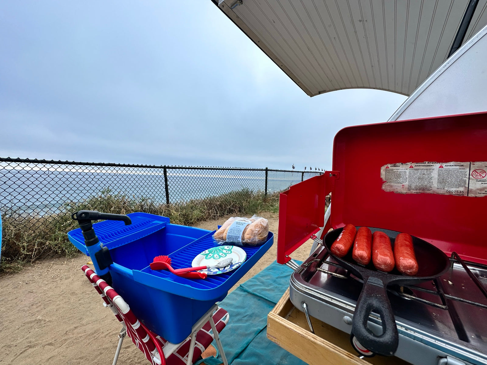 Outdoor cooking by the ocean: Hot dogs sizzle on a camp stove, set up outside an RV rental, offering scenic views.