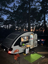 Twilight camping with a modern rv rental teardrop trailer, solar panels & open kitchen in a serene pine forest setting.