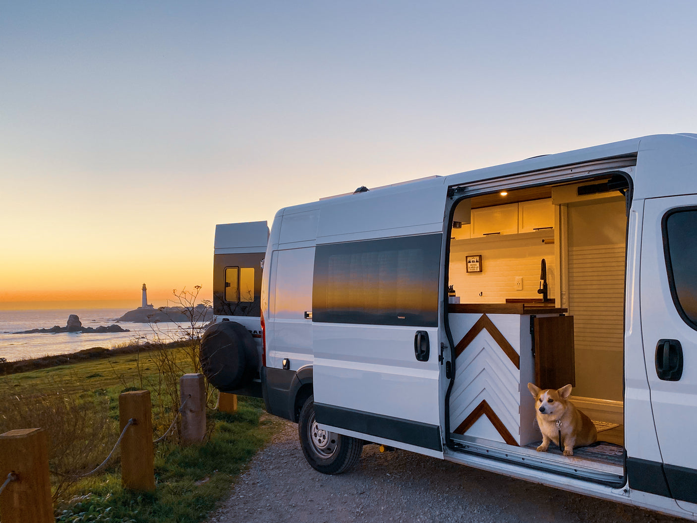 Coastal RV rental adventure with a Corgi, enjoying a vibrant sunset by a lighthouse. The cozy van interior glows warmly.