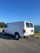White Ford cargo van parked on a coastal road overlooking the Pacific Ocean. Perfect for an RV rental getaway.