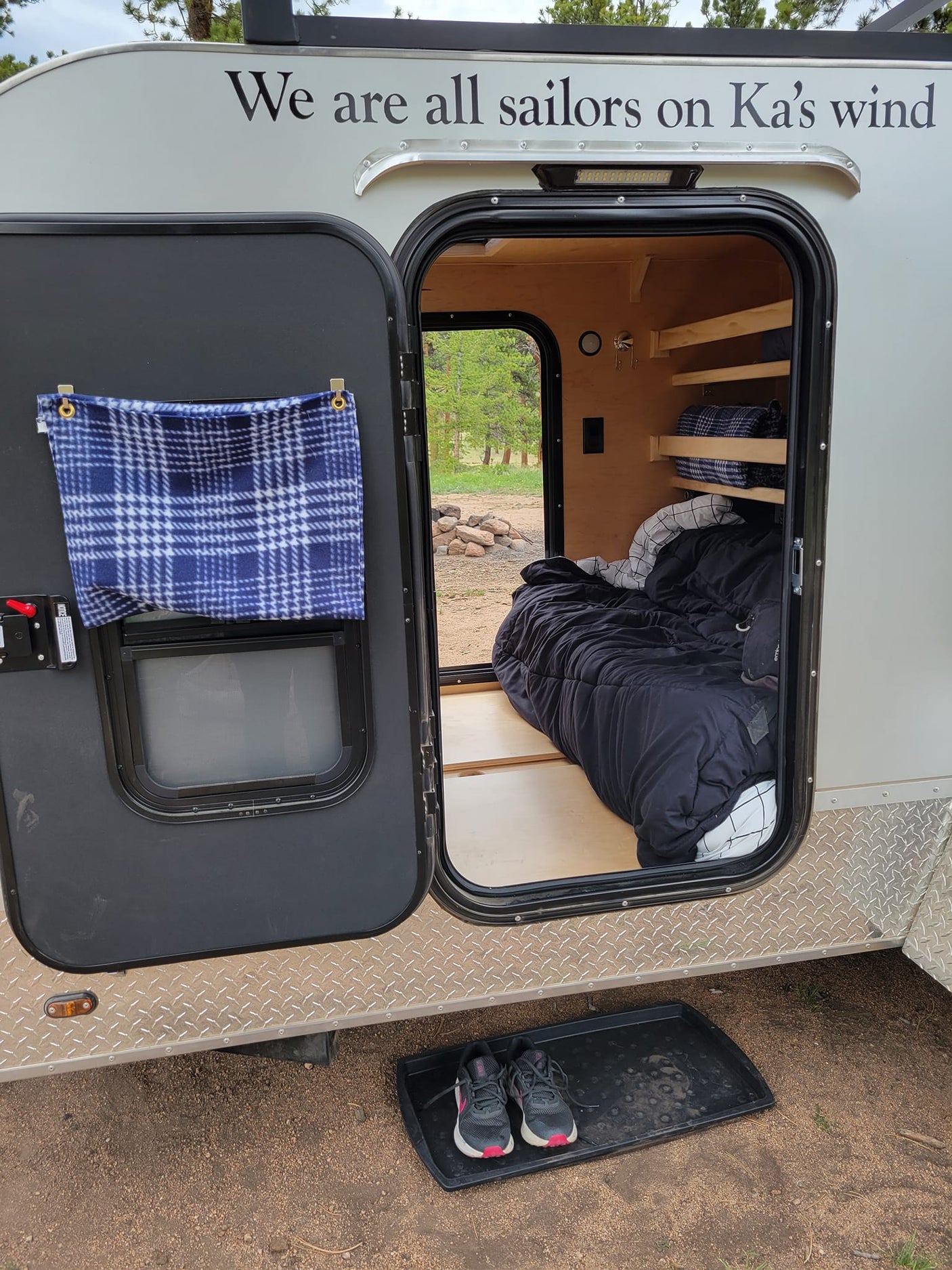 This cozy RV rental teardrop trailer features an open door, revealing a comfy bed & wooden shelves inside. Perfect for camping, shoes outside.